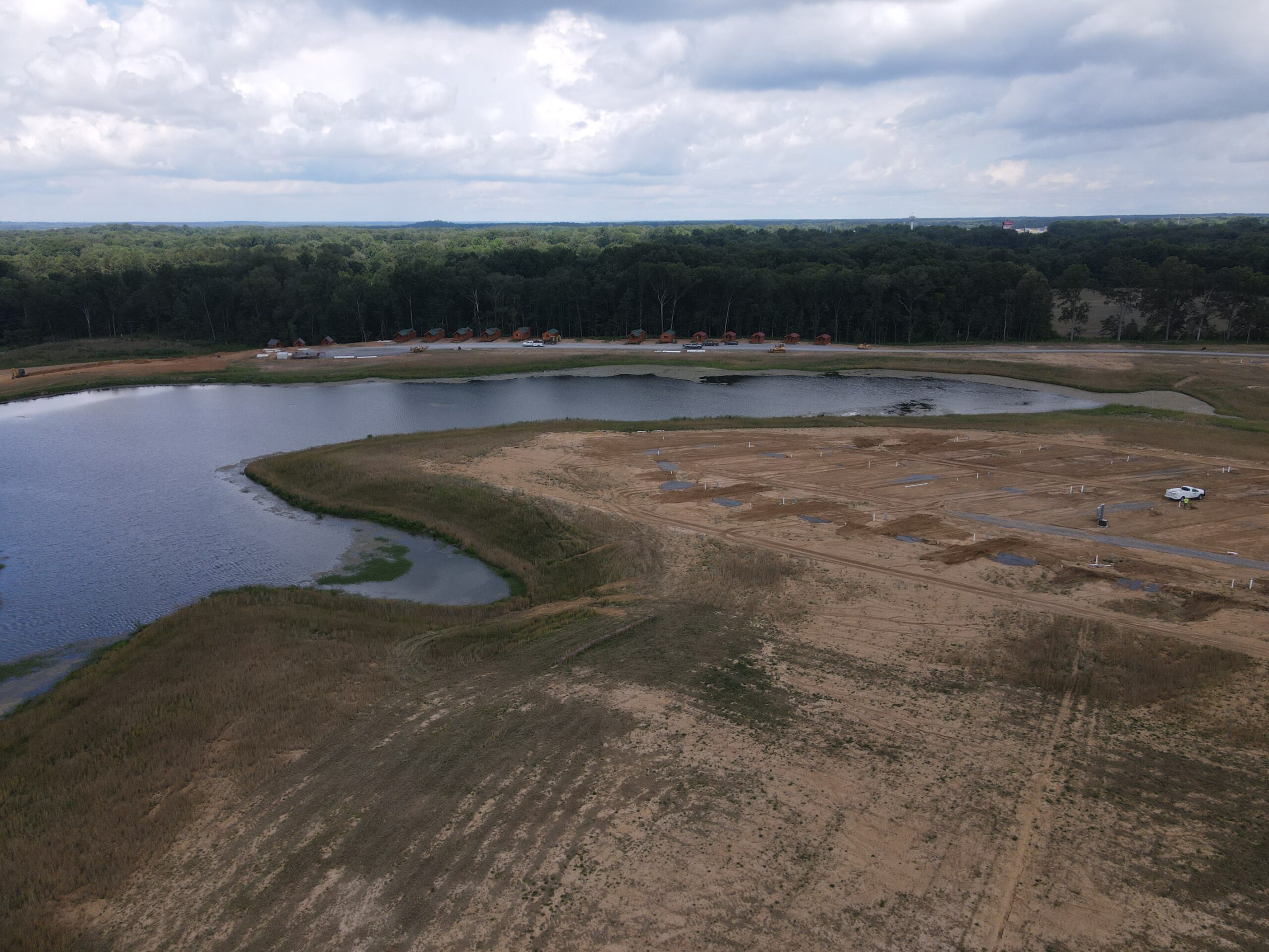 Aerial view of a construction site near a lake with patches of bare earth, grassy areas, a few vehicles, and small cabins lined along the far side of the lake, bordered by dense trees under a cloudy sky.