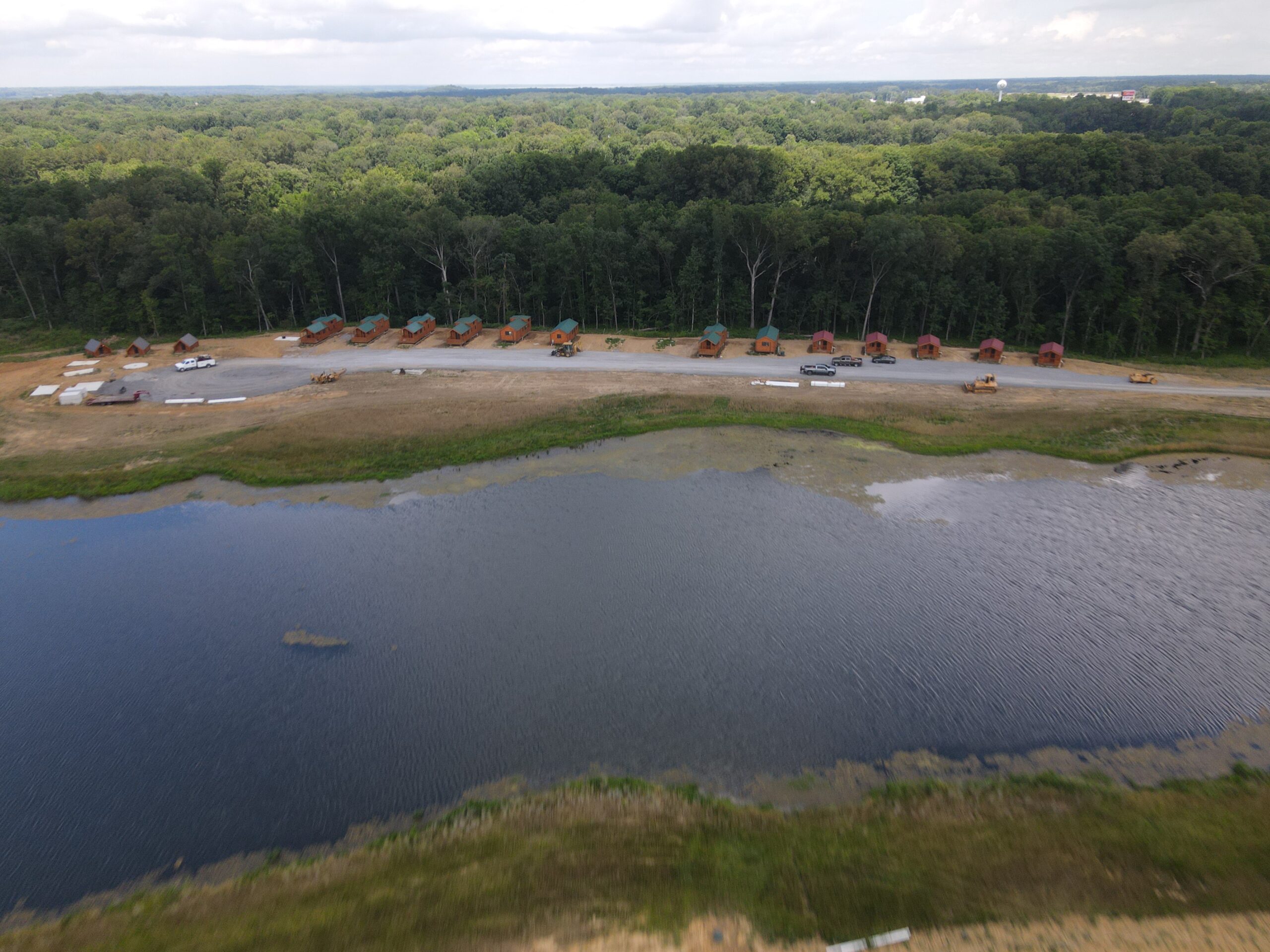 A row of construction vehicles and equipment is lined up on a dirt road beside a wooded area, with a body of water in the foreground and dense trees in the background under a cloudy sky.