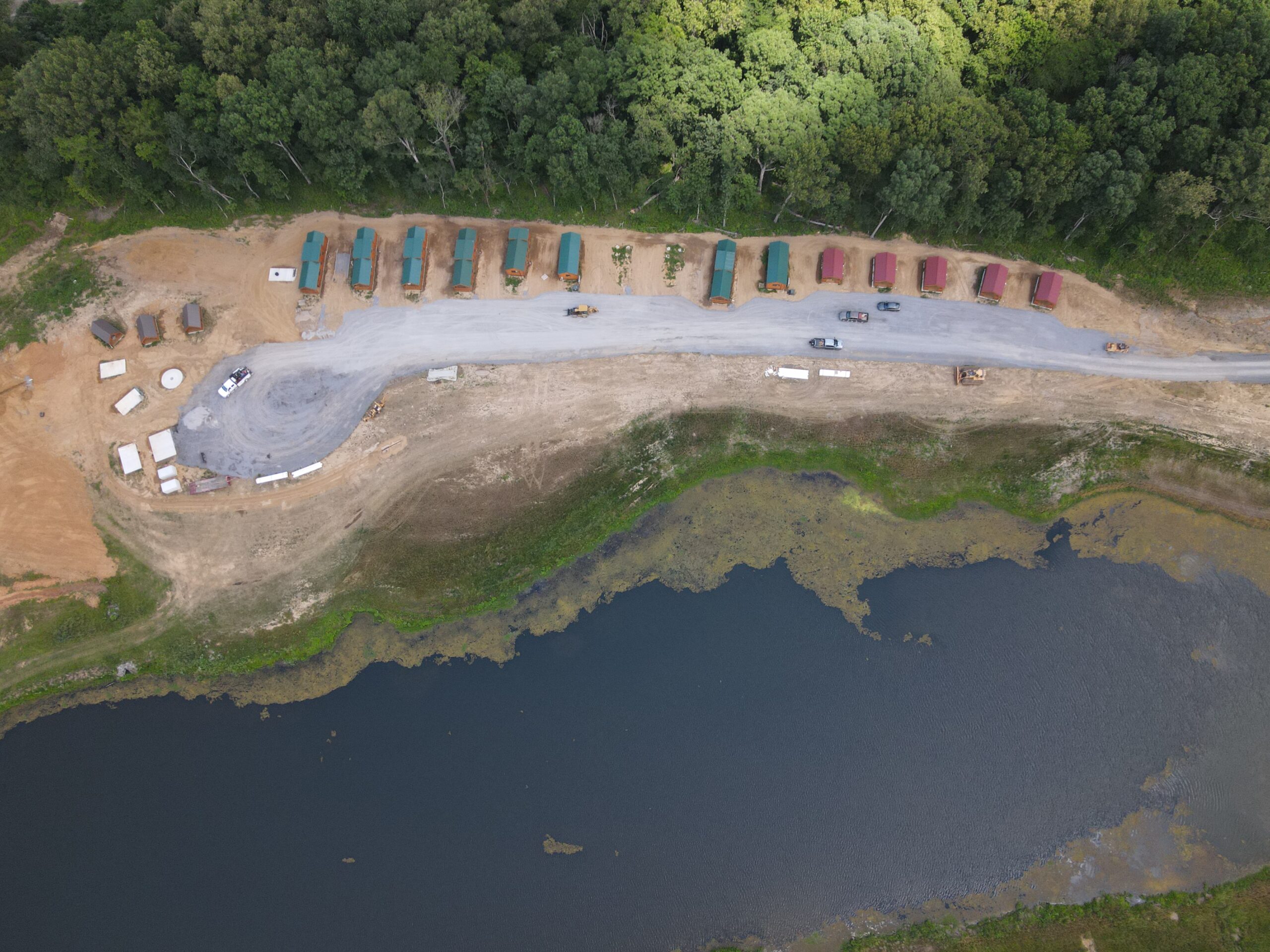 Aerial view of a construction site near a forest, with rows of cabins or containers lined along a road, several vehicles parked nearby, and a pond bordered by grass at the bottom of the image.